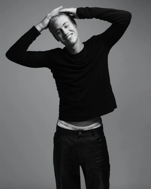 Black and white portrait of a smiling young adult man in dark clothing with his hands on his head in a photo studio setting.