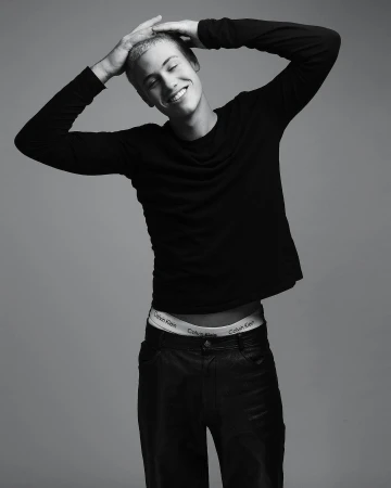 Young man smiling with hands behind his head wearing a black long-sleeve shirt and dark pants against a grey background