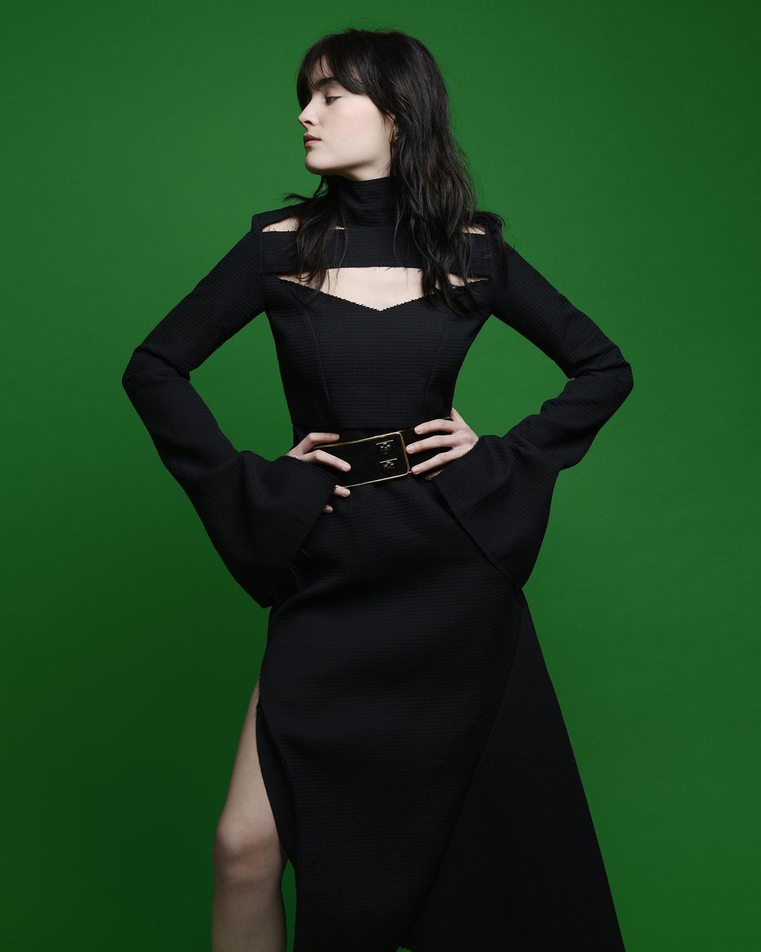 A brunette woman models a black cut-out dress with a slit, accessorized with a wide gold belt, against a vibrant green background.