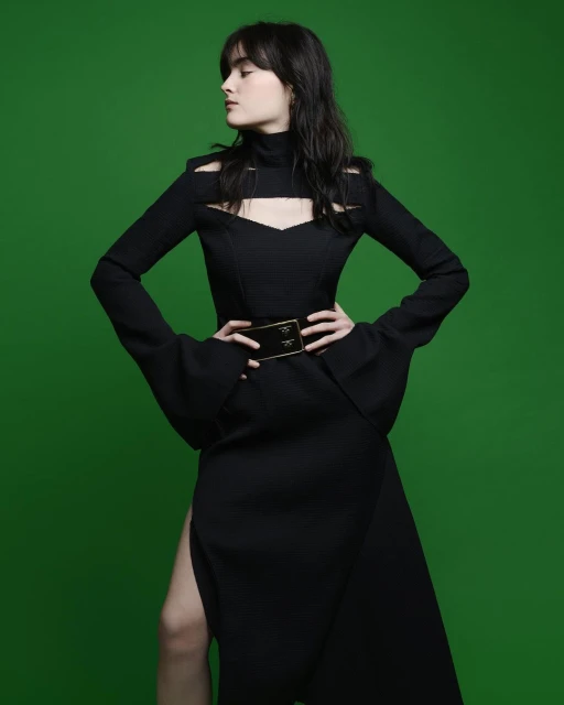 A brunette woman models a black cut-out dress with a slit, accessorized with a wide gold belt, against a vibrant green background.