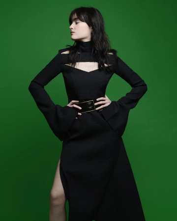 A brunette woman models a black cut-out dress with a slit, accessorized with a wide gold belt, against a vibrant green background.