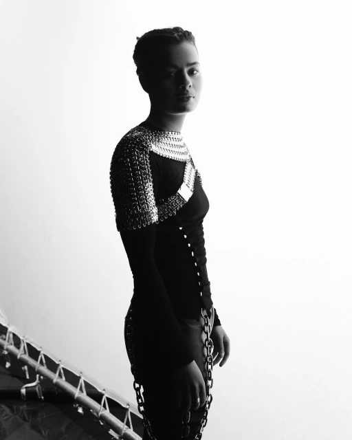 Black and white portrait of a young woman in a fashionable metal chainmail top and chains, striking a confident pose against a white backdrop.