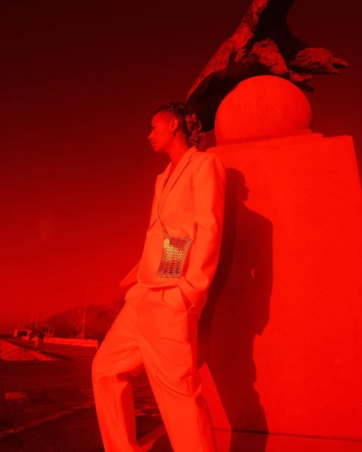 An adult leaning on a red plinth with sculpture, wearing a matching suit and a small metallic bag, with an outdoor, red-tinted ambiance.