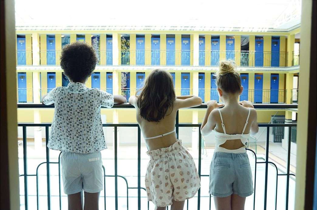 Three children stand on a balcony overlooking a building with rows of blue and yellow doors, providing a rear view of the kids.