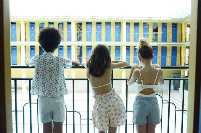 Three children stand on a balcony overlooking a building with rows of blue and yellow doors, providing a rear view of the kids. 