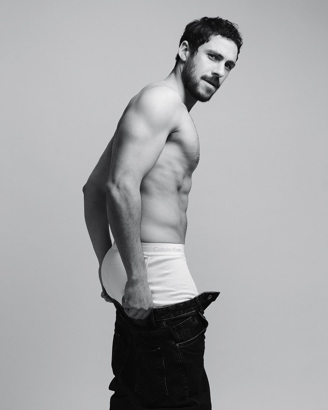 Black and white portrait of a shirtless man in underwear and jeans, looking back over his shoulder against a gray backdrop.