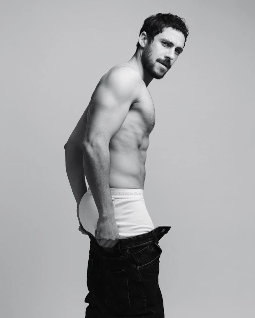 Black and white portrait of a shirtless man in underwear and jeans, looking back over his shoulder against a gray backdrop.