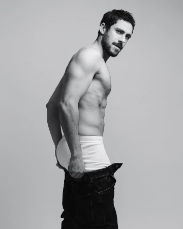 Black and white portrait of a shirtless man in underwear and jeans, looking back over his shoulder against a gray backdrop.