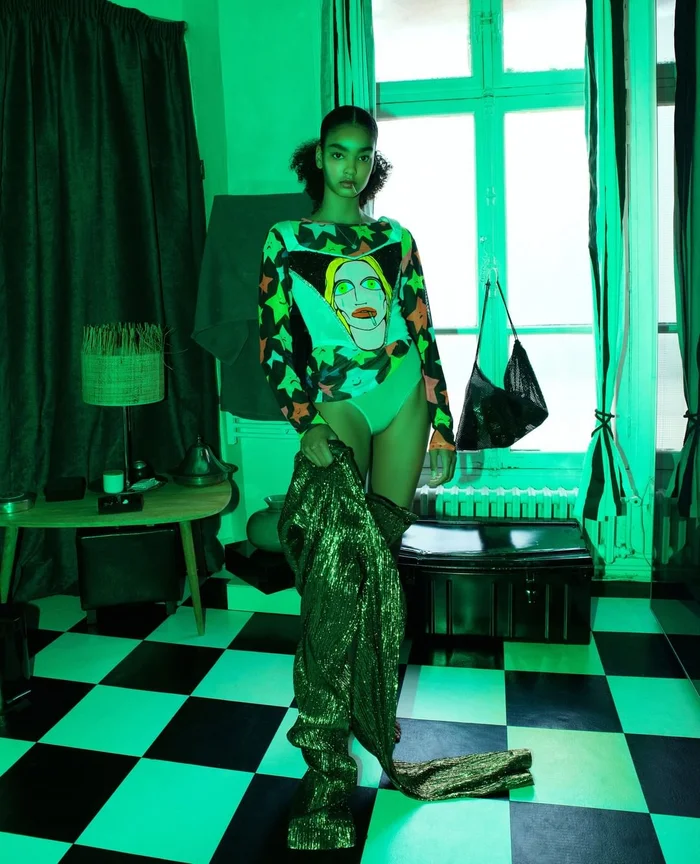 Model standing in a green lit room wearing a graphic printed top and holding a textured metallic garment.