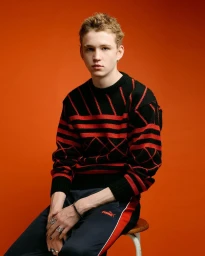 Young man in a black and red patterned sweater and dark trousers seated against a solid orange studio backdrop.