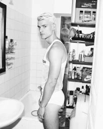 Blonde man in a white tank top standing in a tiled bathroom, photographed in black and white by Matthieu Dortomb.