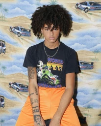 Young man with dark curly hair wearing a black graphic t-shirt and orange pants against a patterned background.