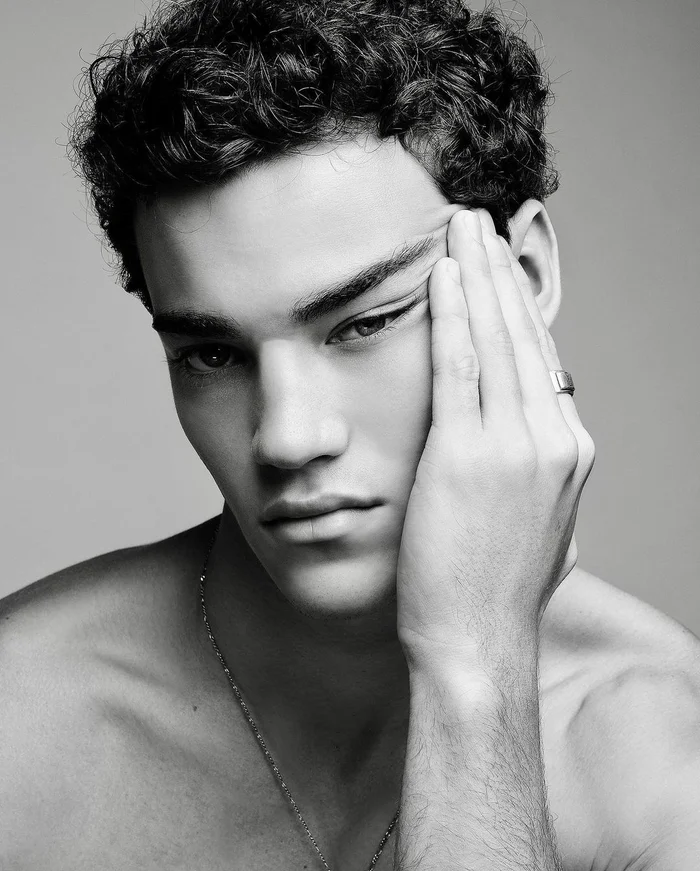 Portrait of young man with dark curly hair touching his temple against plain grey backdrop