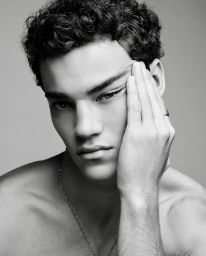 Portrait of young man with dark curly hair touching his temple against plain grey backdrop
