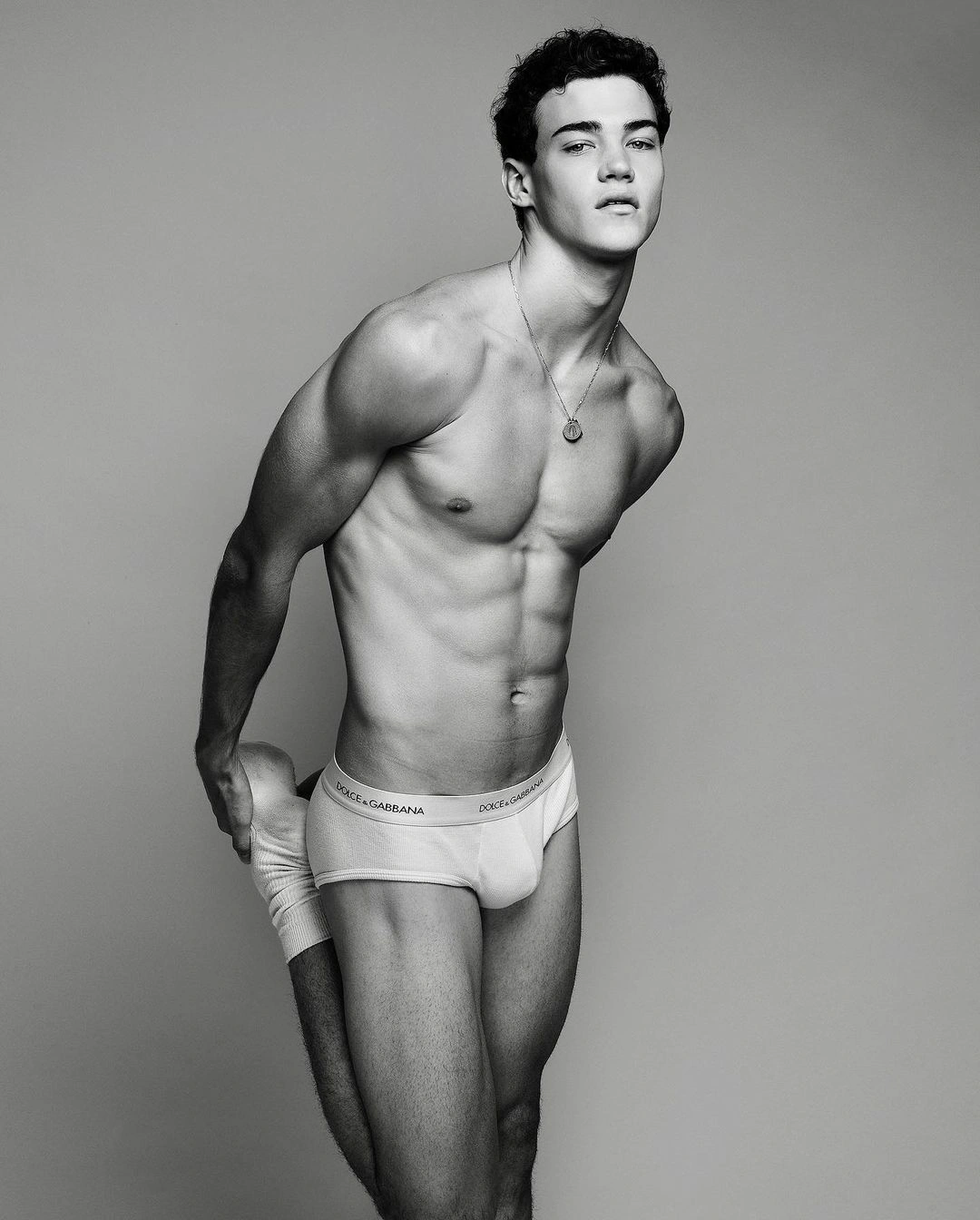 Black and white portrait of a male model wearing branded briefs while stretching against a plain grey studio backdrop