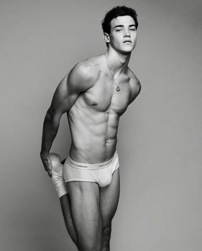 Black and white portrait of a male model wearing branded briefs while stretching against a plain grey studio backdrop