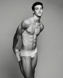 Black and white portrait of a male model wearing branded briefs while stretching against a plain grey studio backdrop