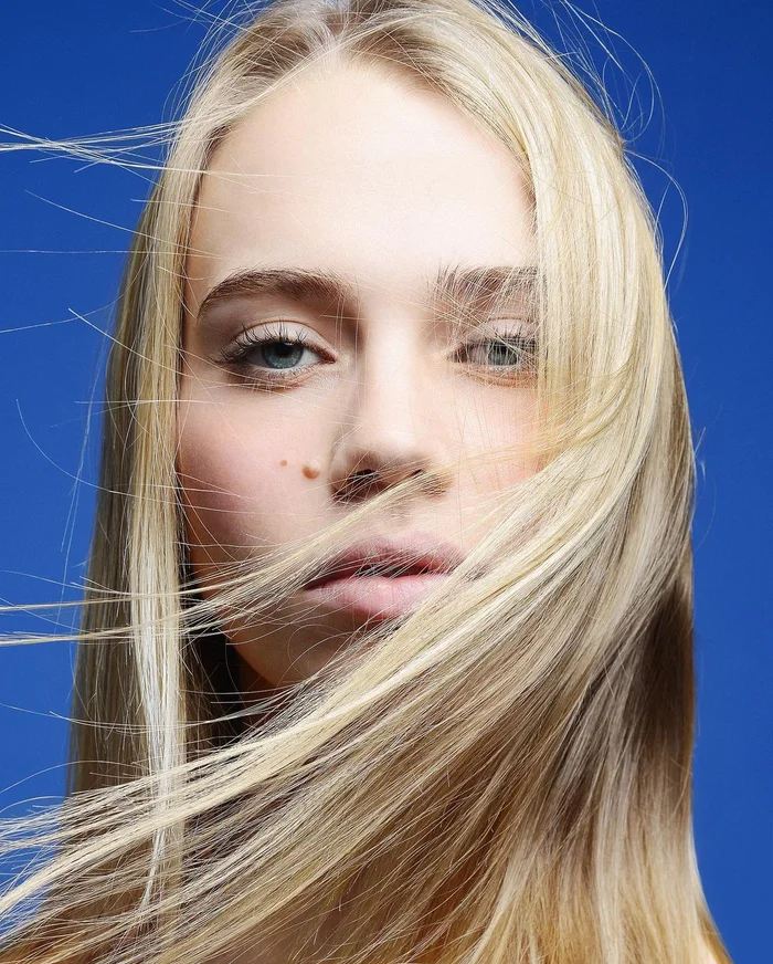 Close up portrait of a blonde woman with flowing hair across her face against a bright blue background