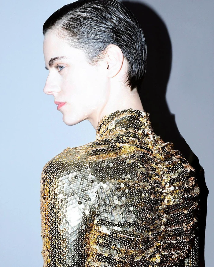 Profile portrait of a woman wearing a sparkling gold sequined high-neck top against a grey wall with a sharp shadow.