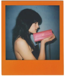 A polaroid style portrait of a woman holding a pink crossbody bag against a plain wall.