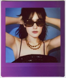 Woman wearing tortoiseshell sunglasses and a gold chain necklace in a portrait captured on instant film with a metallic purple border.
