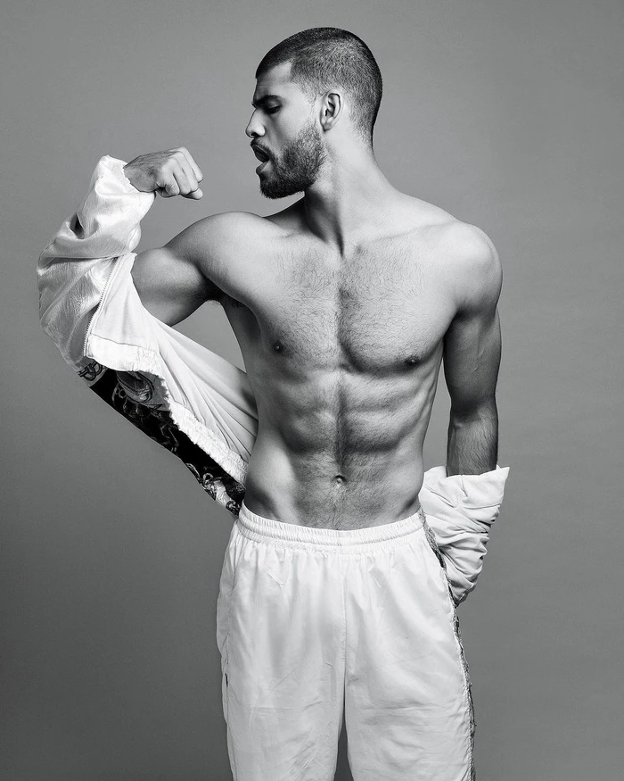 Monochrome portrait of a muscular shirtless man flexing his bicep while wearing white athletic trousers.