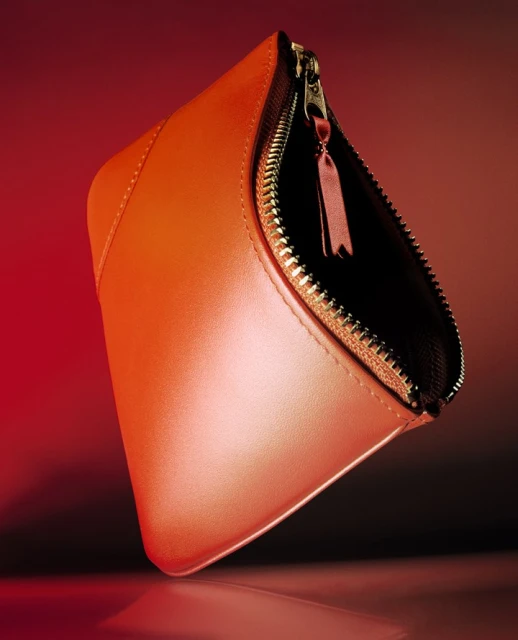 Close-up of a coral-colored cosmetic bag with a zipper on a reflective surface, set against a gradient red backdrop.