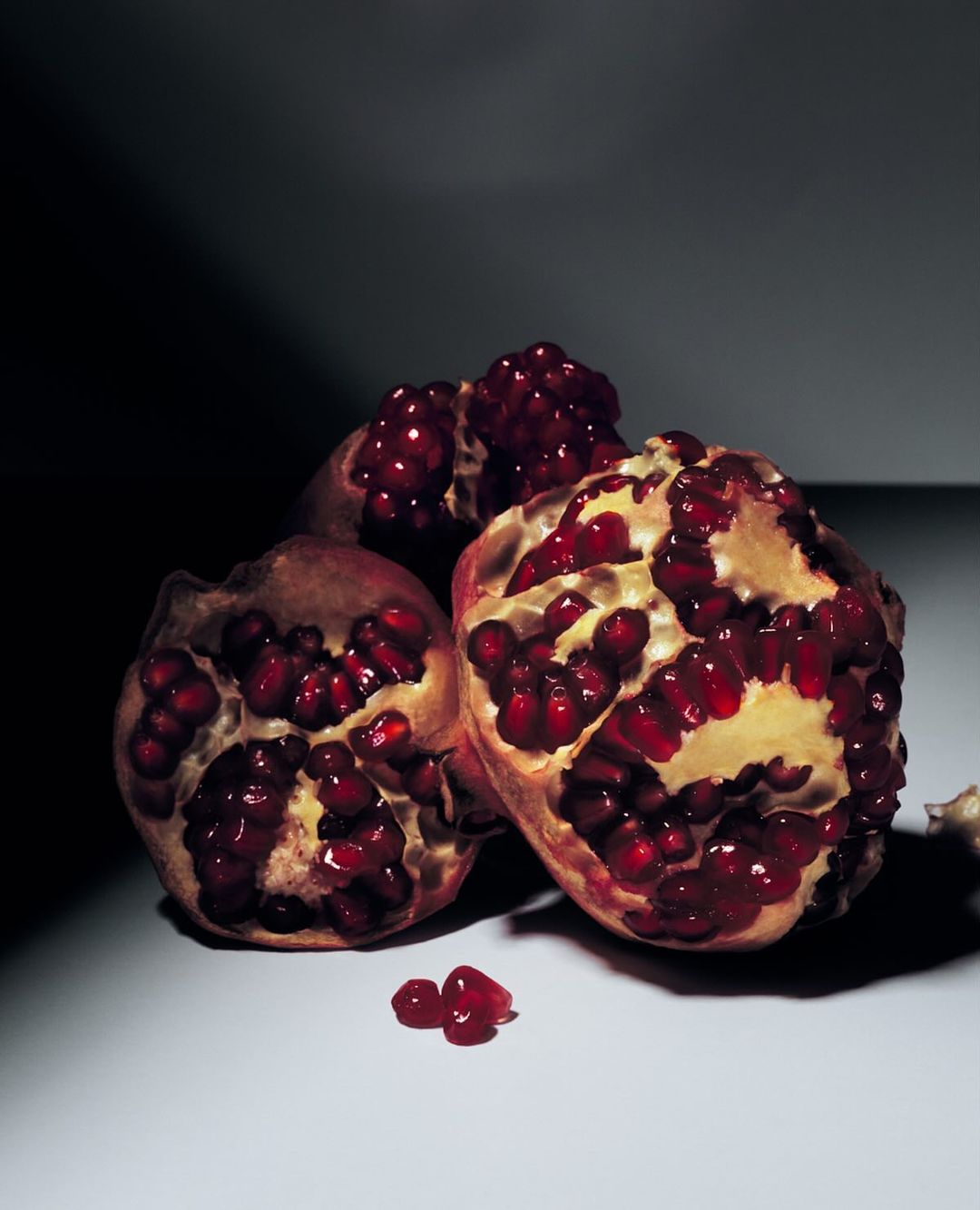 Close-up of three pomegranate halves with red seeds on a white surface against a dark background, studio lighting emphasizes texture.