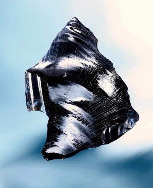 Striking studio shot of a shard of black obsidian glass, its surface reflecting light in swirling patterns, set against a soft blue gradient backdrop.