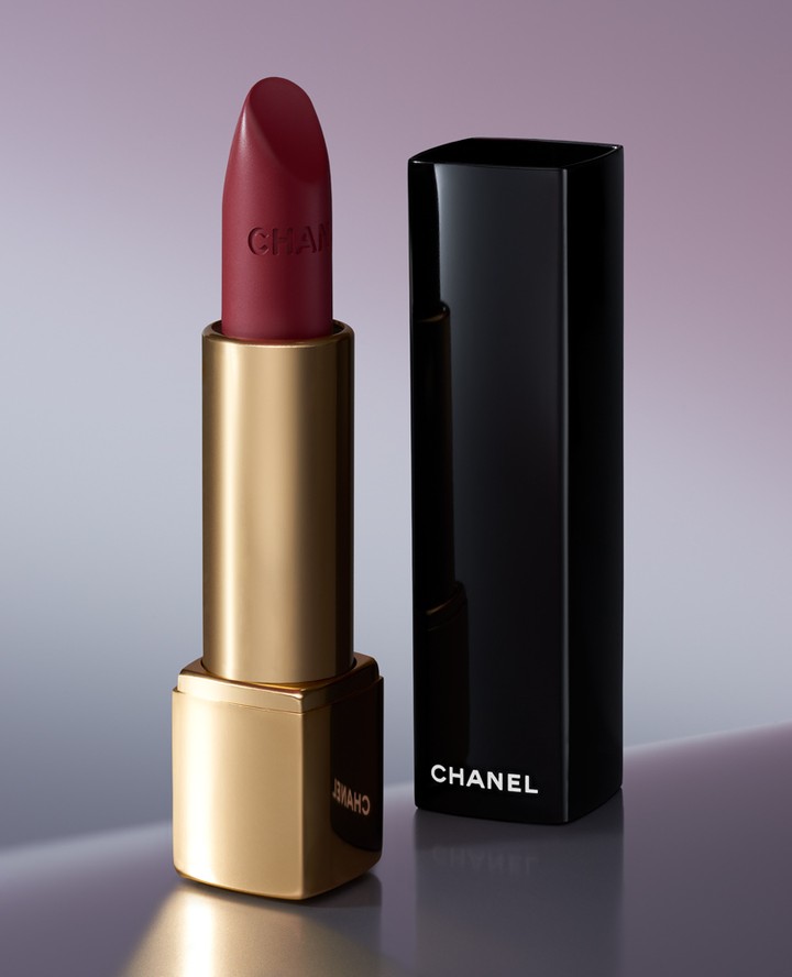 Close-up of a red Chanel lipstick tube with gold base and black cap on a softly lit, neutral gradient background. Beauty shot.