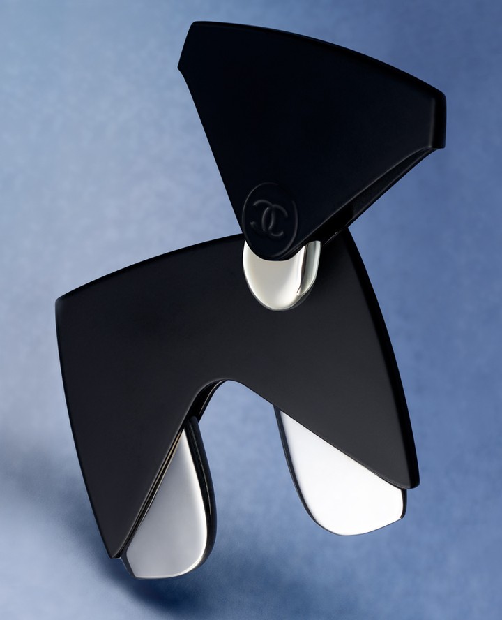 Stylish black and silver brooch featuring the iconic Chanel logo, set against a soft blue backdrop, perfect for luxury fashion accessory.