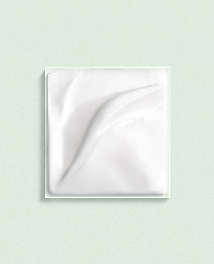 Elevated studio shot showcases a white cream sample in a clear, square container against a solid light green backdrop, focusing on texture.