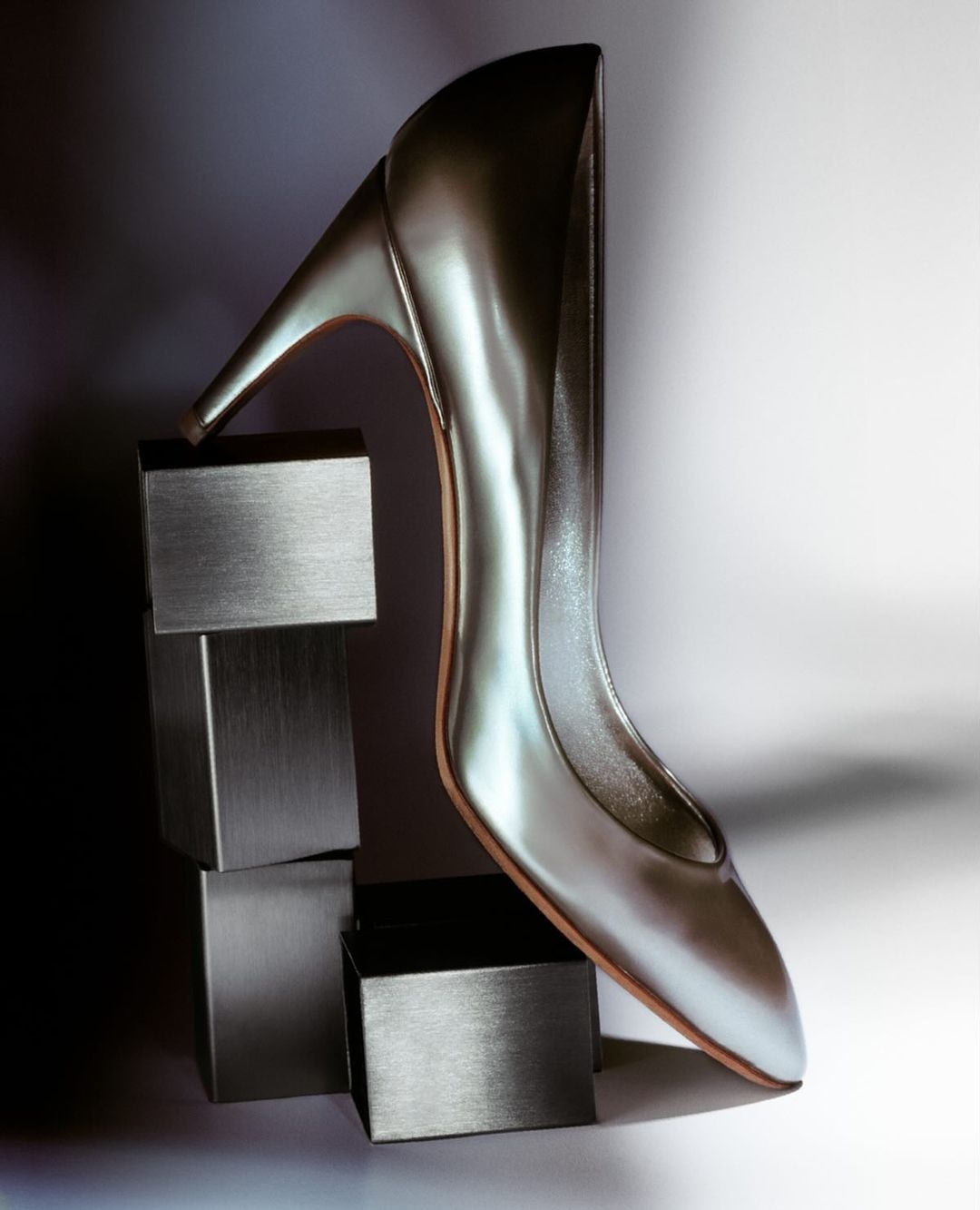 Elegant, metallic silver high-heel shoe is elevated by stack of metallic silver cubes, with a bright, graduated white background.