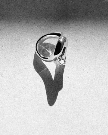 A sculptural silver ring with a single embedded gemstone resting on a textured grey background with a sharp shadow.