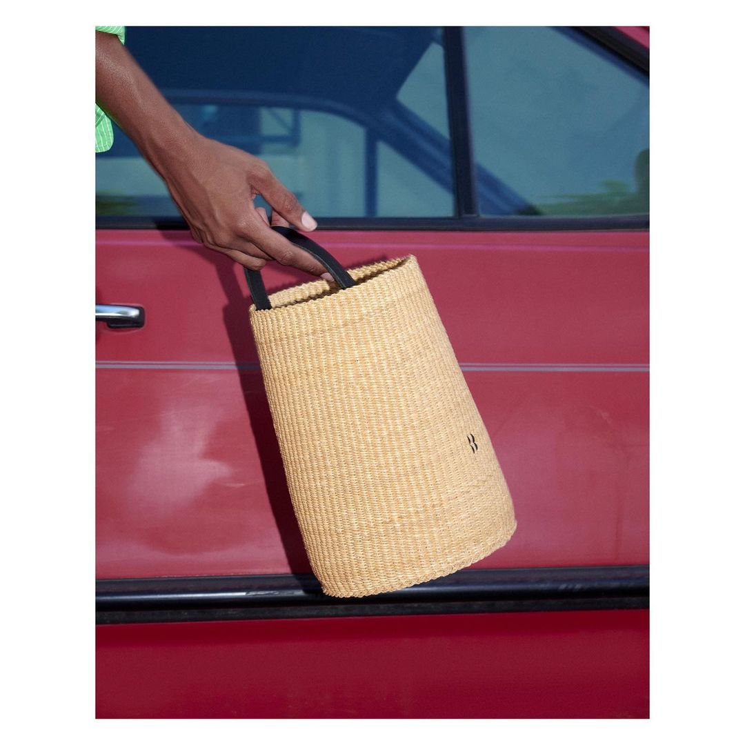 Close-up of a person holding a woven handbag with black handles against a red car door, showcasing fashion and style.