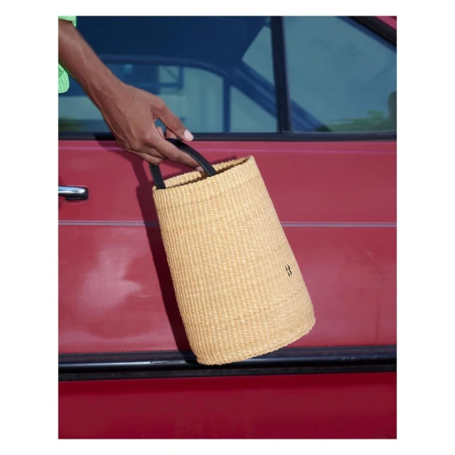 Close-up of a person holding a woven handbag with black handles against a red car door, showcasing fashion and style.