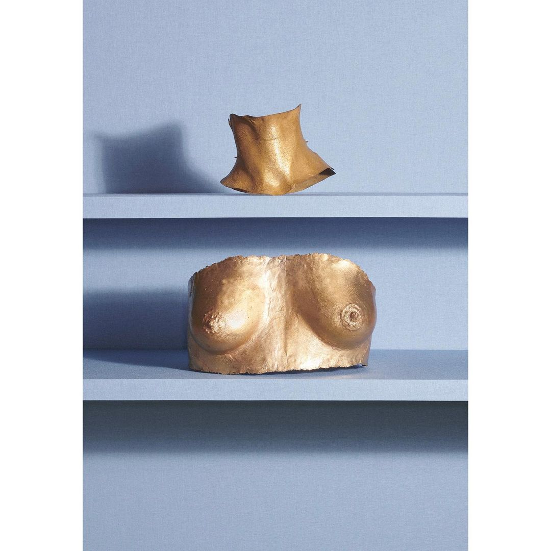 Golden sculpted forms of a bust and neck rest on a blue tiered shelf, presented in a conceptual art studio photograph.