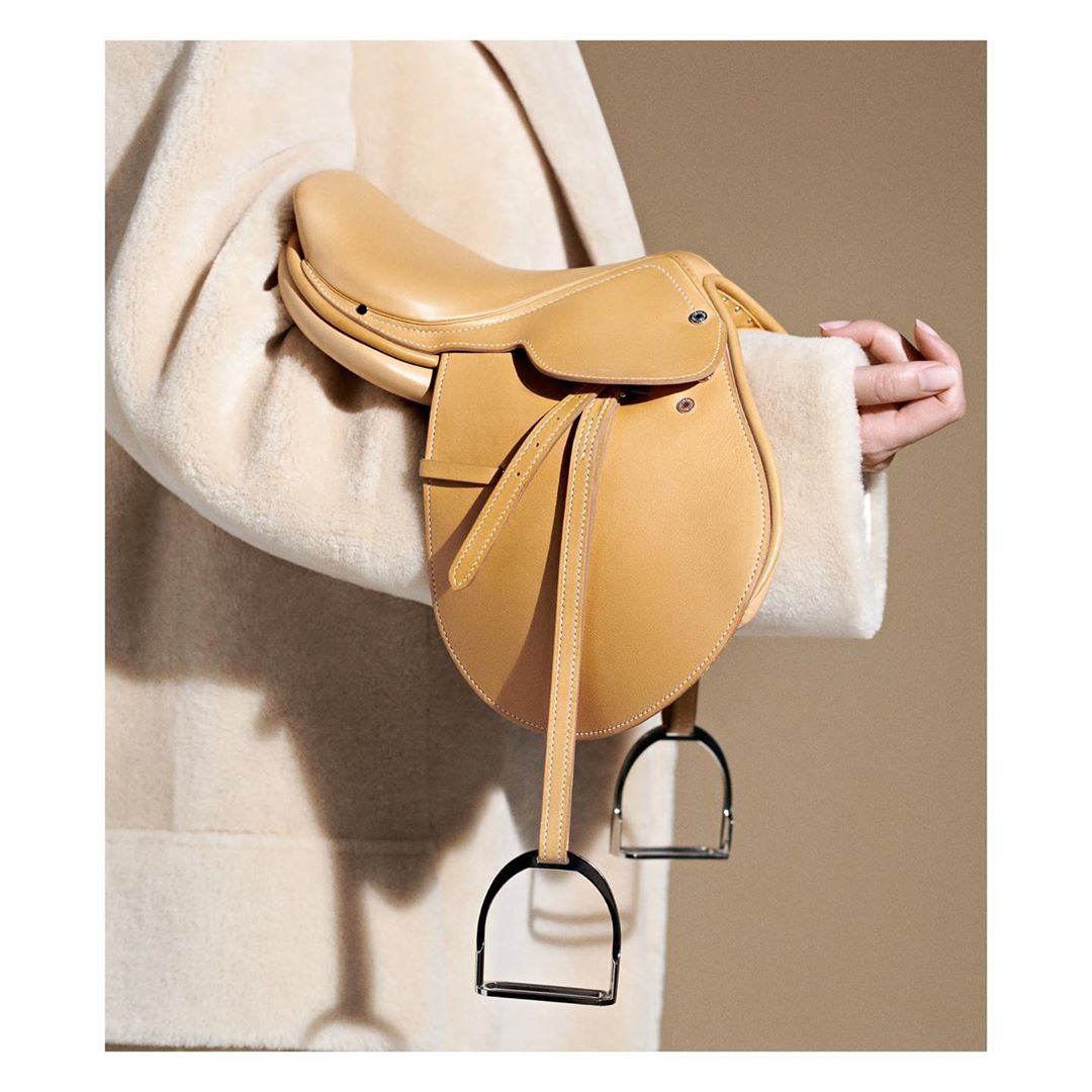 Equestrian saddle purse held by a person in a coat, in a studio setting with soft, neutral lighting and a tan background.