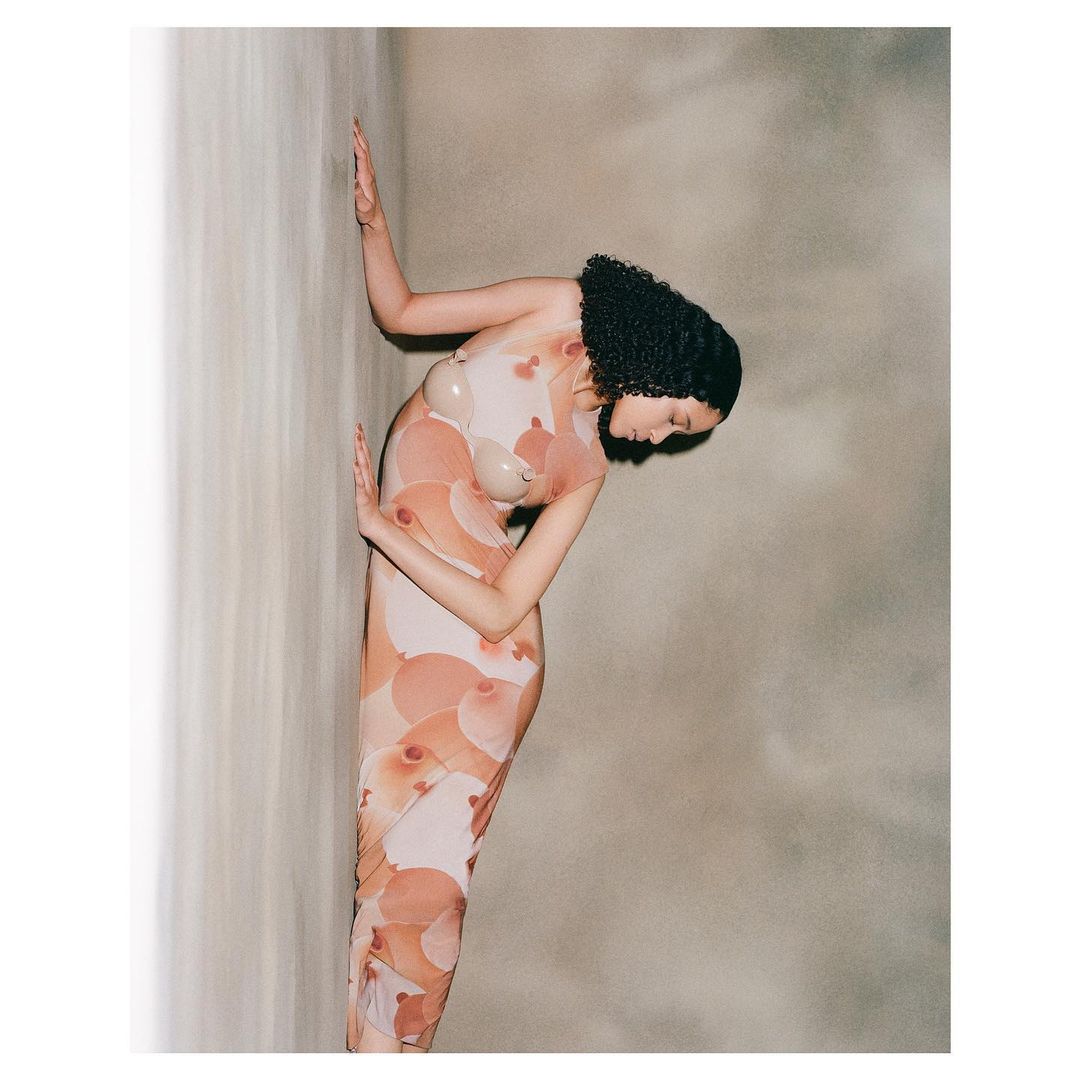 Woman in nude-toned dress with floral pattern leaning against a wall in a studio setting. Dark curly hair and neutral background.