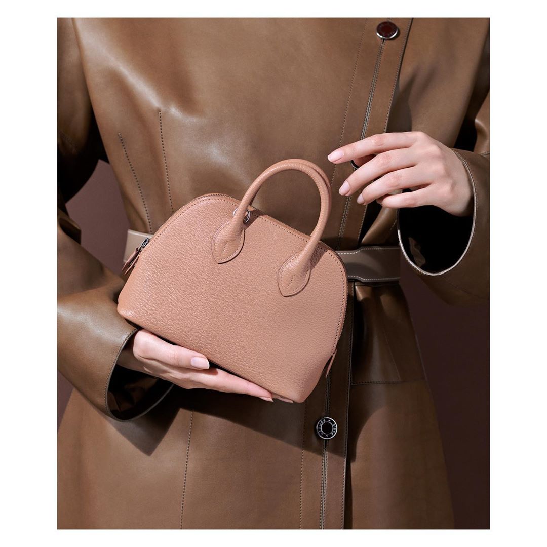 Tan-colored dome handbag held by a person wearing a tan leather coat, showcasing fashion accessory and style in a studio setting.