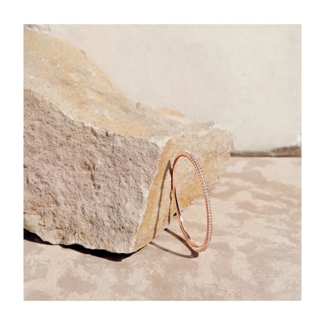 Close-up of a sleek rose gold bangle with gemstone detailing, resting against a textured stone on a neutral-toned surface.