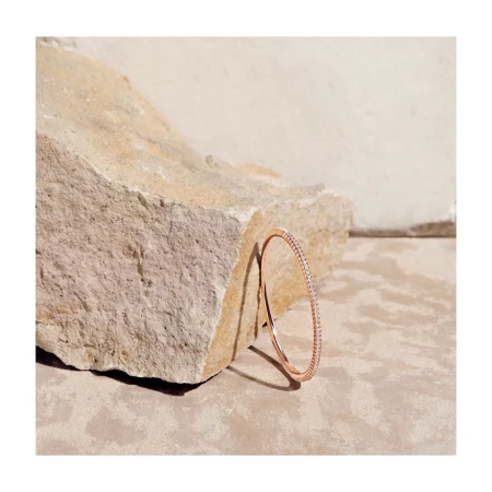 Close-up of a sleek rose gold bangle with gemstone detailing, resting against a textured stone on a neutral-toned surface.