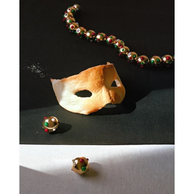 Still life of a masquerade mask with gold beaded jewelry on a divided black and white surface under bright light creating strong shadows.