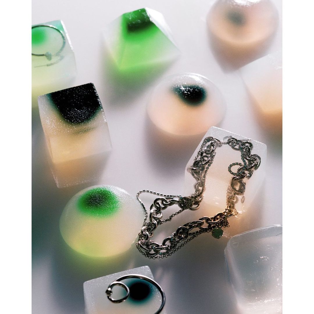 Studio shot of a silver chain necklace frozen inside an ice cube, surrounded by other colorful ice cubes against a white background.