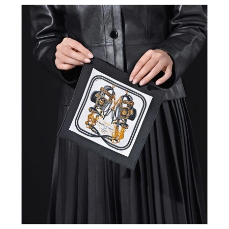 Woman holds a black clutch featuring a printed scarf panel with equestrian details, wearing a black leather jacket and pleated skirt.