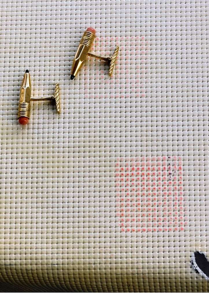 Close-up of a pair of gold pencil-shaped cufflinks with coral erasers displayed on a textured white fabric background with a red pattern.