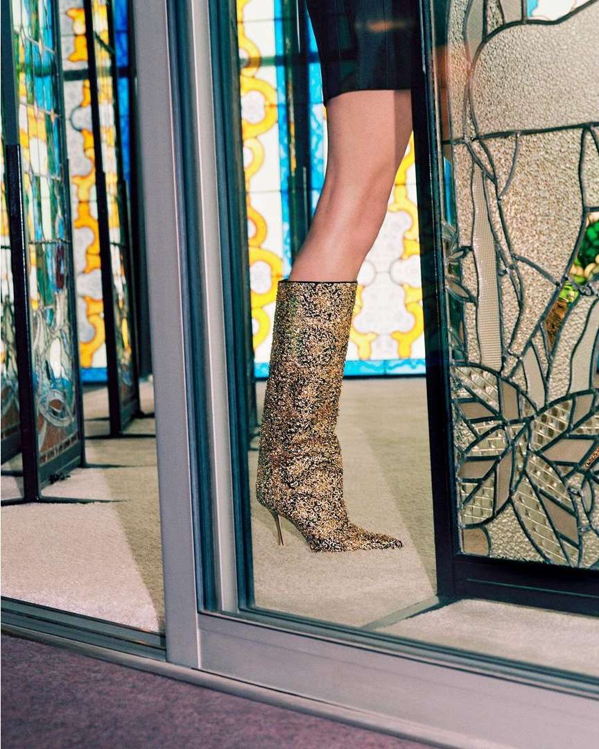 Fashionable sparkly gold stiletto boot is reflected in glass panels creating an infinite view in an indoor setting on carpet.