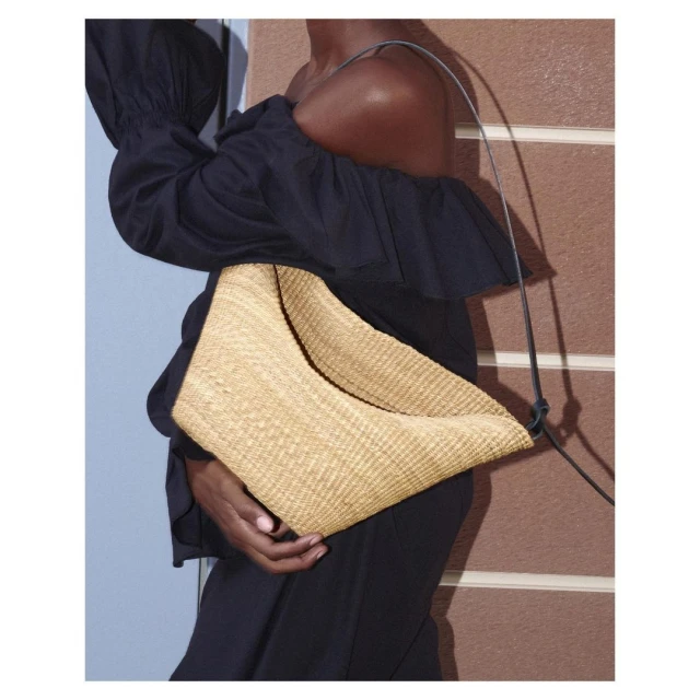 Studio shot of a dark-skinned woman in a black off-shoulder dress, modeling with a woven straw-textured bag on a neutral background.