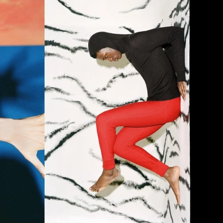 Fashion image of a person in a black hooded shirt and red pants lying against a wall with a black and white abstract pattern.