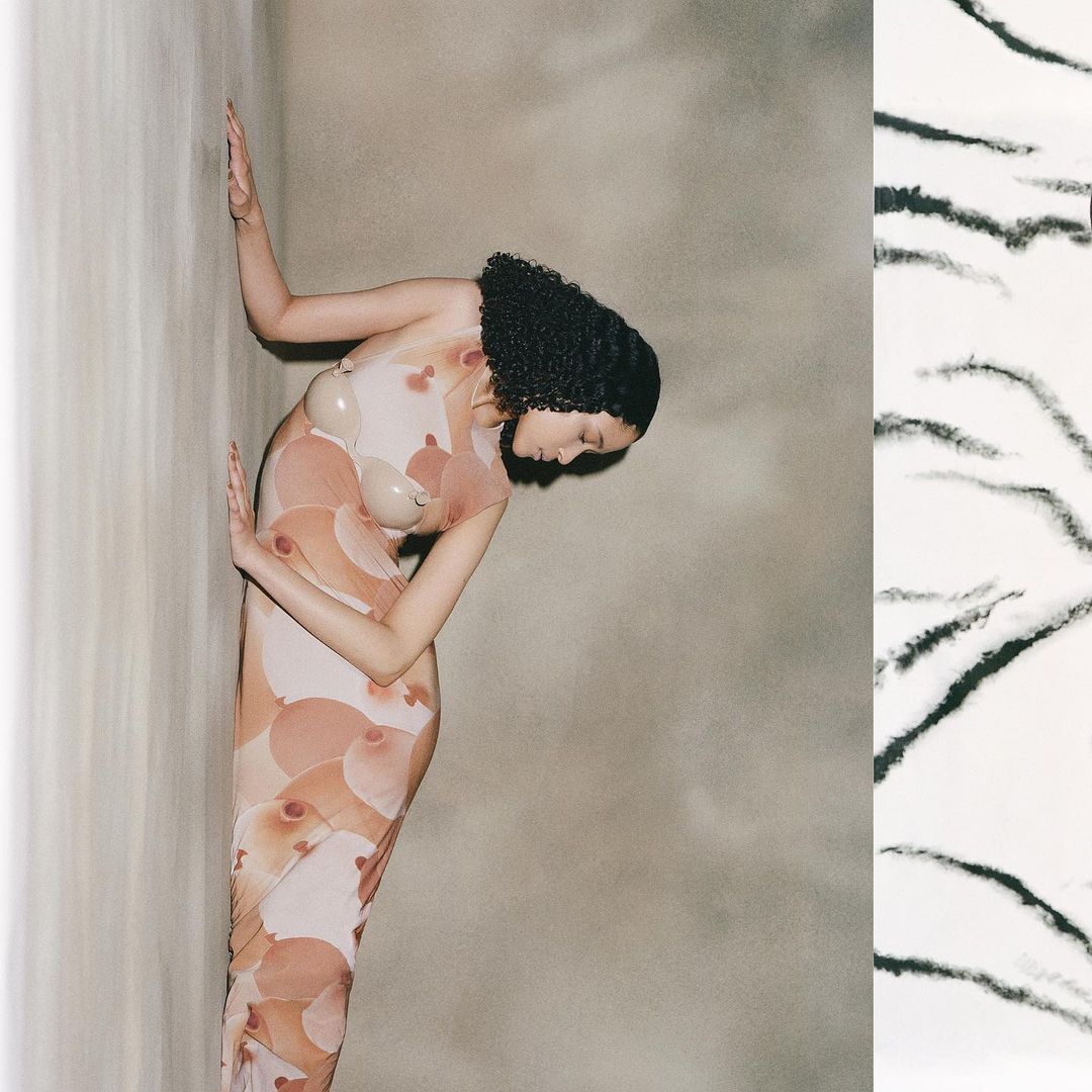 Elegant woman with dark, curly hair poses in a nude illusion dress with breast-shaped appliques in a neutral-toned studio setting.